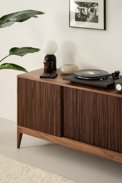 A modern interior scene featuring a wooden sideboard with vertical grooves, topped with a variety of items. To the left, there is a green potted plant. On the sideboard surface are a black and white table lamp, a candle, a rectangular object, a small decorative bowl, and a record player with a vinyl record. Above the sideboard, a monochromatic abstract art piece is mounted on the white wall. The floor is light and adorned with a cream-colored carpet. Record Player Stand Ideas, Record Player Corner, Manly Living Room Ideas, Music Corner Living Room, Record Credenza, Vinyl Player Stand, Vinyl Record Table, Vinyl Cabinet, Vinyl Area