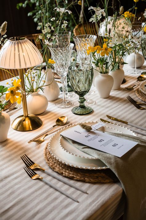 Striped tablecloth with wicker place mat with white plates, sage green napkins and monochrome wedding menu stationery with white and gold cutlery including green coloured glassware with small lamps and wildflowers in white vases | wedding table decor | green and yellow wedding colour palette | Jacqui McSweeny Wedding Green Table Decor, Sage Napkins Place Settings, Sage Green Wedding Tablescape, Green Glasses Wedding, Sage Green White And Yellow Wedding, Yellow Green White Color Palette, Green Plates Wedding, Green Yellow White Wedding, White And Yellow Wedding Theme