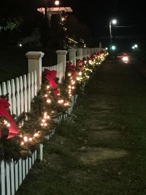 Picket Fence Christmas garland Picket Fence Holiday Decor, Fence Decor For Christmas, Christmas Lights On Rod Iron Fence, Wood Fence Christmas Decorations, Christmas Garland Fence Ideas, White Picket Fence Christmas Decor, Wooden Fence Christmas Decorating Ideas, Fence Decorations Christmas, White Fence Christmas Decor