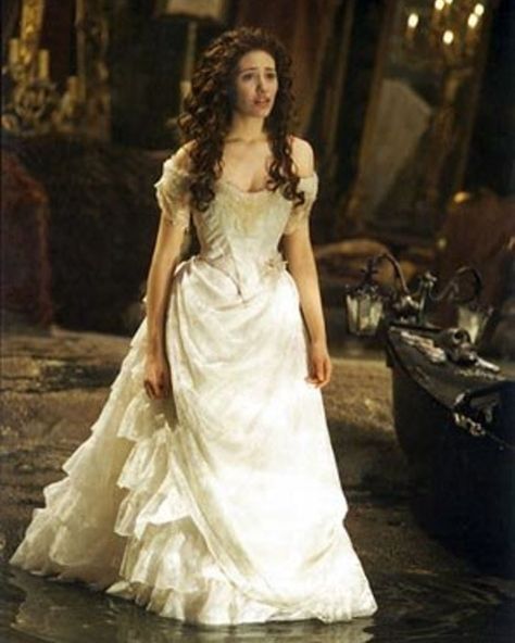 "The Phantom of the Opera" Wedding Dress  As creepy as we find the Phantom's obsession with Christine (Emmy Rossum) in the 2004 film adaptation of the Broadway musical, you have to admit: The masked man sure can dress a hostage bride. Made by costume designer Alexandra Byrne, the off-the-shoulder, lace-trimmed ivory ball gown is a symbol of the character's virtue. It sold  for $5,120. Phantom Of The Opera Wedding, Opera Wedding, Halloween Wedding Dresses, Movie Wedding Dresses, Ivory Ball Gown, Opera Dress, Wedding Dress Costume, Christine Daae, Iconic Weddings