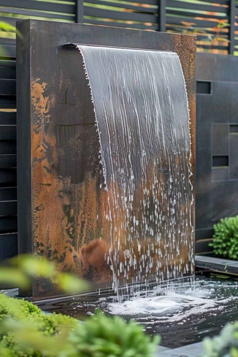 A rustic metal wall fountain with water gracefully flowing over a textured surface, surrounded by greenery in a stylish garden setting. Metal Fountains Outdoor, Waterfall From Wall, Outdoor Patio Fountain Ideas, Water Feature For Patio, Outdoor Wall Fountains Modern, Waterfall In Backyard, Metal Water Feature, Waterfall Water Feature, Water Features In The Garden Fountain