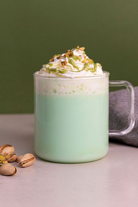 A green coloured pistachio and rose flavoured hot chocolate, made with white chocolate powder. The drink is in a transparent mug, sat on a grey table next to some pistachios, all against a dark green background. The drink is topped with whipped cream, pistachio flavoured sauce and crushed pistachios. Pistachio White Hot Chocolate, Pistachio Hot Chocolate, Romantic Drinks, Pistachio Syrup, White Hot Chocolate Recipe, Chocolate Coffee Beans, Milkshake Machine, Coffee Syrups, Slushie Recipe