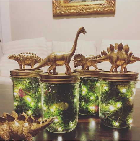 Mason jar centerpieces, lights ,eggs , gold glitter,  and dried moss Jar With Lights Centerpiece, Dinasour Centerpiece, Dinosaur Flower Centerpiece, Dino Theme Centerpiece, Dino Themed Centerpieces, Green And Gold Dinosaur Party, Dino Party Centerpiece Diy, Jurassic Centerpieces, Jurassic World Centerpieces Party Ideas