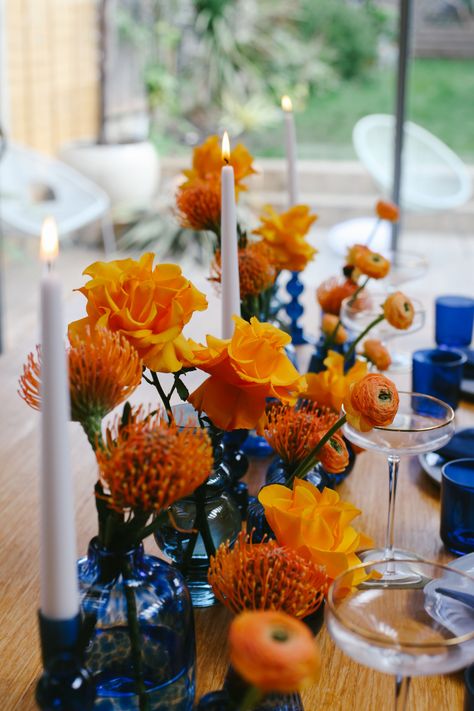 TablescapeBristol Blue GlassOrange table design Orange Blue Table Decor, Table Blue Decoration, Orange And Blue Party Ideas, Orange And Yellow Table Setting, Blue And Orange Centerpieces Wedding, Blue And Orange Event Decor, Navy And Orange Tablescape, Cobalt Blue Table Setting, Navy And Orange Table Setting