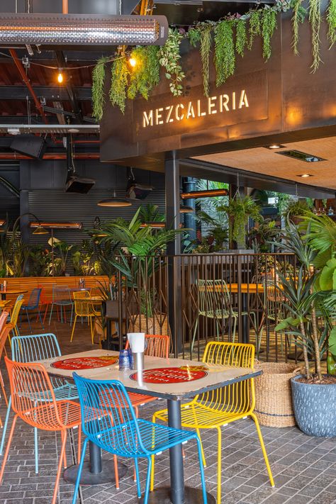 bright coloured seating area with square glossy wooden table, floral background with a mixture of different plants creating a beautiful atmosphere. big open plan restaurant and bar area with black walls and stone floor. Mexican Restaurant Outdoor Patio, High End Mexican Restaurant, Mexican Taqueria Design, Aesthetic Mexican Restaurant, Mexican Cafe Design, Mexican Restaurant Patio, Modern Mexican Restaurant Design, Mexican Bar Ideas, Mexican Restaurant Interior Design