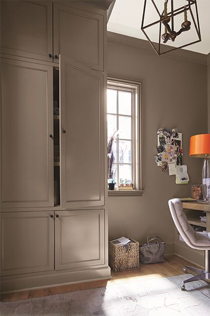 A taupe-painted home office with cupboard, beige chair, wooden desk, note board, white ceiling, and abstract chandelier. Dark Beige Kitchen Walls, Dellwood Sand Benjamin Moore, Best Dark Beige Paint Color, Benjamin Moore Fossil Paint Color, Built In Bookcase Color Ideas, Furniture For Dark Walls, Rich Taupe Paint Color, Falcon Brown Benjamin Moore, Taupe Walls And Trim