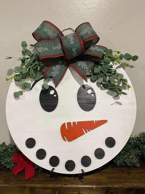 Welcome Winter and your guests with this 18" Snowman Door Hanger. Painted white and lightly weathered, with a green and red bow and a dusty colored greenery for a final touch! - Made From 1/4" Baltic Birch.- Measures 18" x 18" - Ready to hang! *Important Notes* We recommend all signs be hung indoors or in a covered outdoor area to keep your sign protected and beautiful! Processing Time And Shipping Due to demand each sign may take 1-2 weeks to cut, assemble, and package with lots of care. Please Wreath With Snowman, Round Snowman Door Hanger, Welcome Cricut Sign, Fall Front Door Signs Wooden, Circular Christmas Sign, Cricut Winter Door Sign, Circle Door Signs Diy Christmas, Easy Door Hangers Diy Crafts, Winter Door Rounds