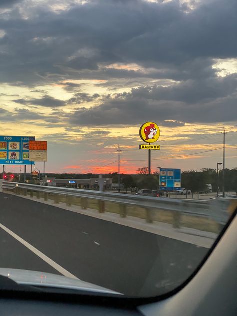 The picture is taken from the passenger seat of a car, looking out over a hot pink sunset far in the distance. In the foreground is a Highway sign listing local places to stop for food at the next exit (Taco Bell, McDonald’s, etc.) and a large sign for the gas station chain Buccee’s Texas Life Aesthetic, Bucees Texas Aesthetic, Buccees Aesthetic, Texas Astethic, Galveston Texas Aesthetic, Texas Summer Aesthetic, Atx Aesthetic, San Antonio Texas Aesthetic, Austin Texas Pictures