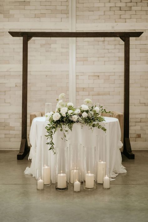 This sweetheart table is decorated with a lush floral arrangement featuring white roses, baby's breath, queen anne's lace, eucalyptus, and italian ruscus. Floral design by Imogen Floral Design. White Florals Sweetheart Table, Sweetheart Table White Flowers And Greenery, Wedding Sweetheart Table With Candles, Sweetheart Table Wedding Candles On Floor, Sweetheart Table Wedding Eucalyptus, Sweetheart Table With Eucalyptus, Minimalist Wedding Sweetheart Table, Head Table Wedding Decorations Candles, White And Green Sweetheart Table Flowers