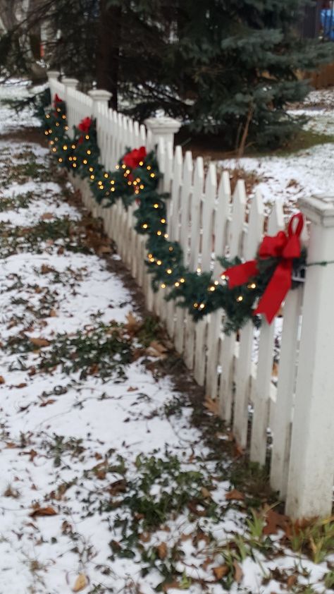 Lighted garland on the picket fence. Garland On White Picket Fence, Fence Decor For Christmas, Outdoor Xmas Garland Ideas, Christmas Decor Fence Ideas, Christmas Lights On Picket Fence, White Picket Fence Christmas Decor, Holiday Fence Decorations, White Fence Christmas Decor, Xmas Fence Decorating Ideas