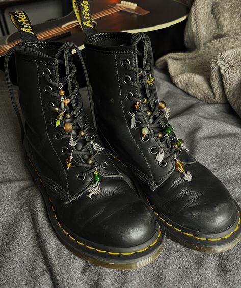 A pair of doc martens with green, brown, and silver charms and beads sitting on grey fabric with a black acoustic guitar in the background Charms On Docs, Diy Doc Martens Charms, Outfit W Doc Martens, Doc Martens With Beads, Decorate Doc Martens, Decorating Doc Martens, Charms On Shoe Laces, Star Shoe Lace Pattern Doc Martens, Doc Martens Beads On Laces