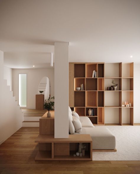 Cozy living space with a soft neutral palette, featuring a large wooden bookshelf as a focal point. A beige sectional sofa is positioned against a low wooden divider, separating the living area from a small staircase leading to another room. The natural wood flooring and minimalist decor, including simple vases and books, create a warm, modern atmosphere, complemented by soft lighting and clean lines. Neutral Minimalist Living Room Cozy, Floating Couch, Wooden Divider, Living Room Minimal, Room Minimal, Sectional Sofa Beige, Small Staircase, Beige Sectional, Modern Living Space