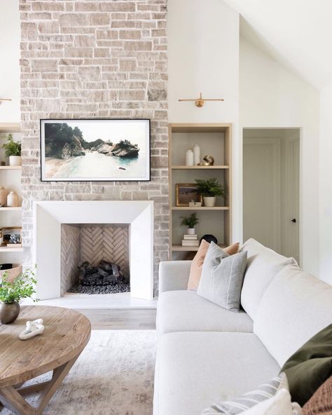 White fireplace with beige herringbone tile firebox is fitted into a floor to ceiling whitewashed stone wall. Black frame beach artwork is mounted above the fireplace in between recessed light wood shelves. Herringbone Fireplace, Beach Fireplace, White Wash Brick Fireplace, White Wash Brick, Shiplap Fireplace, Fireplace Built Ins, White Fireplace, Wood Fireplace, Home Fireplace