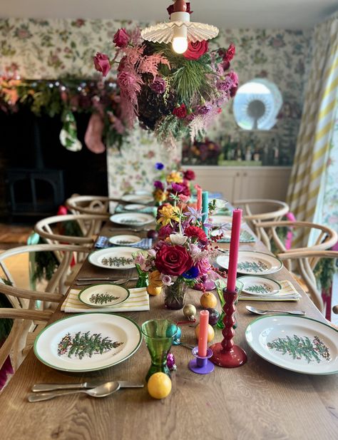 Colourful Christmas tablescape on large wooden table with Spode dinnerware which is white with a green rim and Christmas tree in the centre. Above the table hangs an extravagant foliage cloud filled with greenery, red roses and pink flowers. The centre of the table is fulled with mini floral displays. bright and colourful candlesticks with fruit and baubles scattered throughout. Eclectic Christmas Table Setting, Pink Red Christmas Table, Colourful Christmas Table Setting, Colourful Candlesticks, Colourful Christmas Table, Spode Dinnerware, Modern Christmas Tablescape, Modern Christmas Table, Red And Pink Christmas