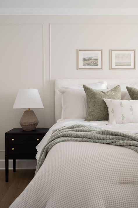 A serene bedroom featuring a neatly made bed with soft, textured white bedding and green accent pillows. A cozy green throw is draped over the bed, adding a touch of nature-inspired color. A black nightstand with gold hardware holds a neutral-toned lamp. Above the bed, two framed landscape artworks hang on the light cream-colored wall, contributing to the peaceful and minimalist aesthetic of the room. The overall atmosphere is calm and inviting, perfect for relaxation. Bedroom With Sage Green Accents, Sage Green Accents, Green Bedrooms, Minimalistic Decor, Natural Vibes, Sage Green Bedroom, Serene Bedroom, Bedroom Retreat, Of Montreal