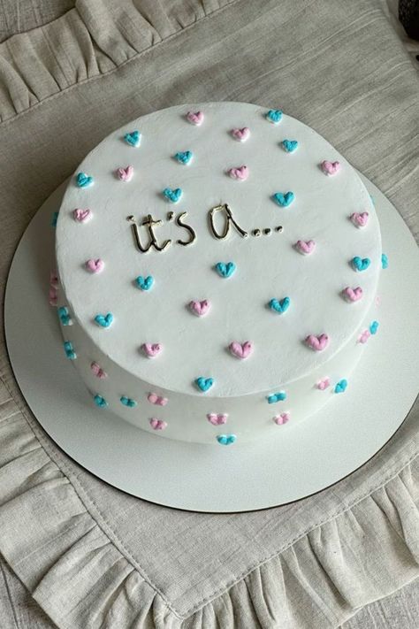 A gender reveal cake with white frosting and pink and blue hearts, with the text "It’s a..." written on top. Simple Reveal Ideas, Gender Reveal Lunchbox Cake, Gender Reveal Cake Diy Easy, Foods For A Gender Reveal Party, Gender Reveal Cake Sayings, Gender Reveal Cutout Cookies, Easy Cute Gender Reveal Ideas, Ways To Do Gender Reveal, Dessert For Gender Reveal Party