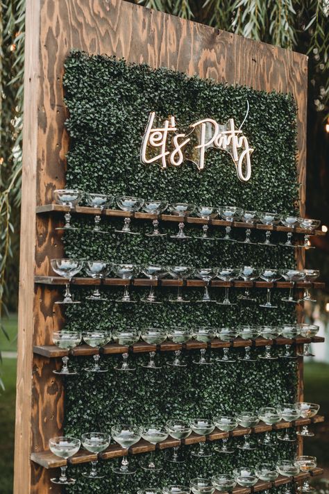 A dark stained walnut champagne wall with wide mouthed champagne glasses, behind it is foliage, at the top there is a LED sign that says "lets party" Wine Wall Display Party, Champagne And Beer Wall Wedding, Beer Tap Wall Wedding, Welcome Champagne Wall, Champagne Flute Wall, Rustic Champagne Wall, Welcome Shots Ideas, Shot Wall Wedding, Wedding Champagne Table