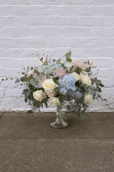 This elegant and gorgeous elevated wedding centerpiece mixes soft white Avalanche roses, pale blue hydrangeas, cream carnations, blush disbud mums, and two kinds of eucalyptus in a lovely silver compote. Blush And Blue Wedding, Carnation Centerpieces, Pale Blue Hydrangea, School Live, Elegant Wedding Centerpiece, Hydrangea Centerpiece, Flower School, Winter Wedding Inspiration, Wedding Centerpiece