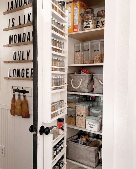 This small but organized pantry optimizes space by putting a spice rack on the door. The shelves are organized with labeled containers and grey fabric baskets. Decorative sign art and a set of small brooms hang near the door. Minimal Pantry, Spice Shelves, Pantry Shelving Ideas, Small Wooden Crates, Black Wire Basket, Wooden Pantry, White Pantry, Small Pantry Organization, Farmhouse Pantry