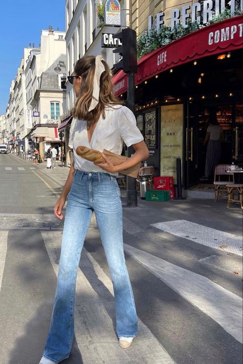 A girl is walking in the middle of the crosswalk in Paris, France. She is wearing light colored highwaisted skinny flared jeans. Her top is a wrap shirt that has a deep-V and it is fully white. Her hair is in a half-up ponytail hairstyle, and a white cream ribbon is tied to the top. Her hair is slightly wavy, and her head is looking to the side with a pair of black sunglasses. Her shoes are white and wicket brown. She is holding a baguette in her hand. The background is a corner coffee shop. Flared Jeans Summer Outfit, Bell Bottom Jeans Outfit Casual, Flare Pants Outfit Summer, Outfits With Bell Bottom Jeans, Bell Bottom Jeans Outfit Summer, Flare Jeans Outfit Summer, Flared Jeans Outfit Summer, Light Denim Jeans Outfit, Flare Jeans Outfit Spring