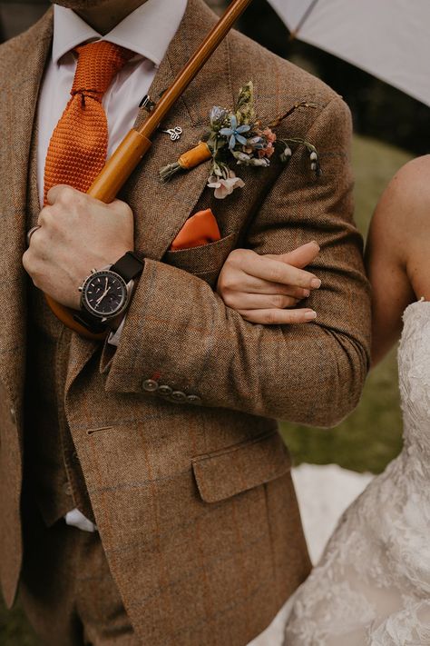 Bride links arms with the groom in brown suit with orange tie and colourful buttonhole with black watch Wedding Suits Groom Fall, Wedding Suits Men Colourful, Colored Suit Groom, October Wedding Suits Men, Coloured Wedding Suit, Fall Wedding Brown Suit, Burnt Orange Groomsmen Suits, Brown Groom Suits For Wedding, Men’s Brown Suit Wedding