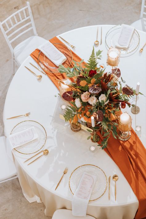 Main wedding table with burnt orange table runner. Decorated with a floral centerpiece alongside candles and furnished with glassware. Wedding Table Decorations Burnt Orange, Burnt Orange Weddings Decorations, Orange Wedding Centerpieces, Orange Wedding Decorations, Orange Centerpieces, Round Wedding Tables, Orange Wedding Themes, Burnt Orange Bridesmaid Dresses, Burnt Orange Wedding