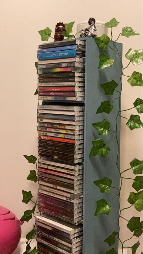 A collection of CDs in a rack covered by fake Ivy. Dvd Shelf Aesthetic, Cd Shelf Aesthetic, Cd Holder Aesthetic, Physical Media Aesthetic, Dvd Organization Ideas, Cd Shelf Ideas, Cd Organization Ideas, Creative Bookshelves For Small Spaces, Cd Display Ideas