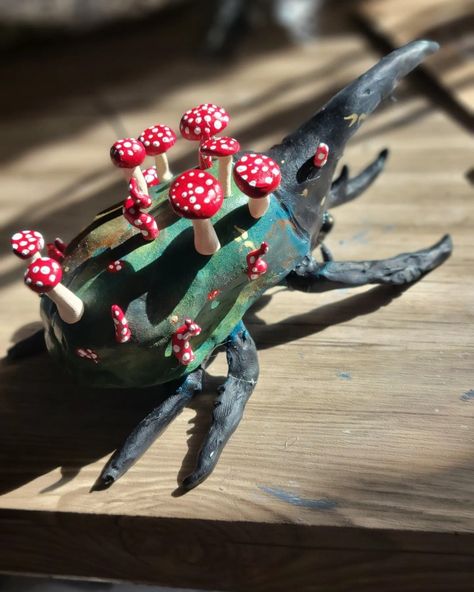 Large beetle with black head, black legs, and green body, facing away from camera, topped with red and white spotted mushrooms. Small gold flecks are scattered across the beetles body, some in the shapes of fae folk wings Paper Mache Beetle, Polymer Clay Beetle, Ceramic Beetle, Bug Ceramic, Clay Beetle, Weird Sculptures, Clay Bugs, Hercules Beetle, Easy Clay Sculptures