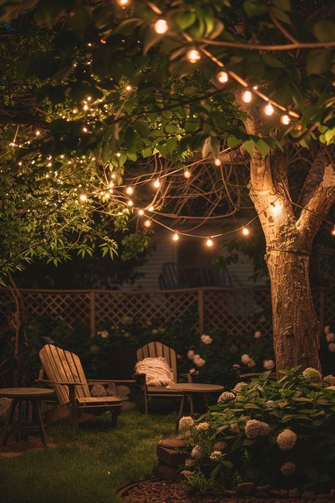 Cozy backyard patio with string lights illuminating wooden chairs surrounded by lush greenery and flowers, creating a perfect outdoor oasis. Twinkle Lights In Trees, Backyard Tree Lighting Ideas, Twinkly Lights Backyard, Fairy Lights Trees Backyards, Lights In Trees Backyard, Ethereal Backyard, Yard Lighting Ideas, Cozy Backyard Ideas, Pretty Backyard