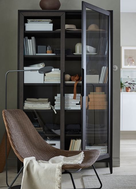 An IKEA REGISSÖR black brown glass door cabinet filled with boxes, books and glassware, behind a chair. Bookshelf Decor With Books, Ikea Regissor, Ikea Glass Door Cabinet, Modern Home Offices, Shelf Cover, Black Bookcase, Bookcase With Glass Doors, Glass Front Cabinets, Ikea Cabinets