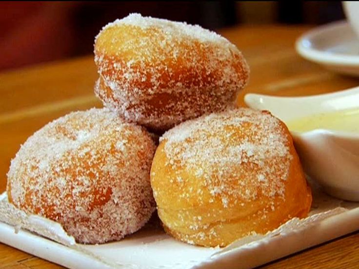 three sugar coated doughnuts on a white plate with dipping sauce in the background