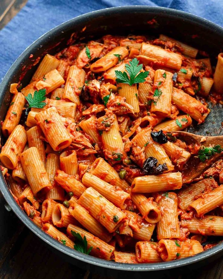 a skillet filled with pasta and sauce on top of a wooden table next to a blue towel