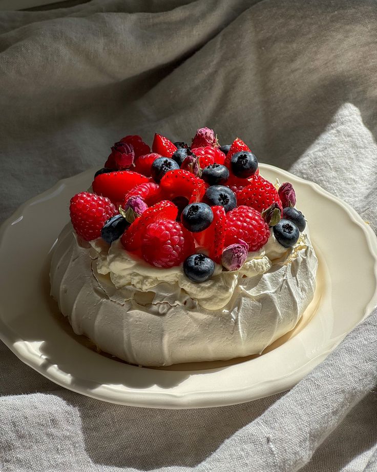 a cake topped with berries and whipped cream on top of a white plate next to a gray cloth