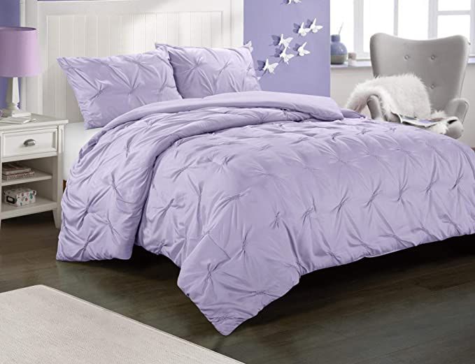 a bed with purple comforter and pillows in a room next to a white chair