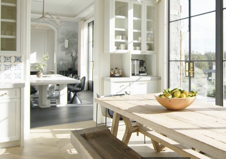 a bowl of fruit sitting on top of a wooden table in front of large windows