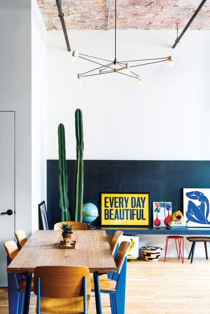 a wooden table surrounded by blue chairs next to a green cactus in a white walled room