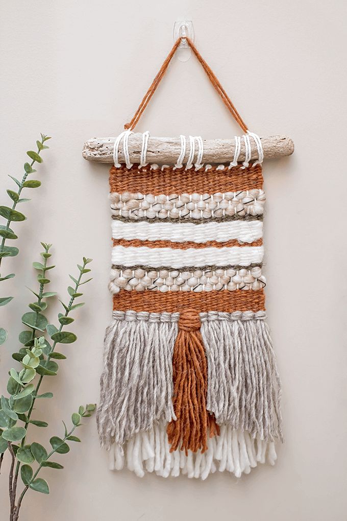 a woven wall hanging with tassels on it next to a potted plant