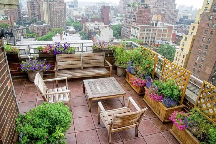 an outdoor patio with wooden furniture and flowers on the roof top, surrounded by tall buildings