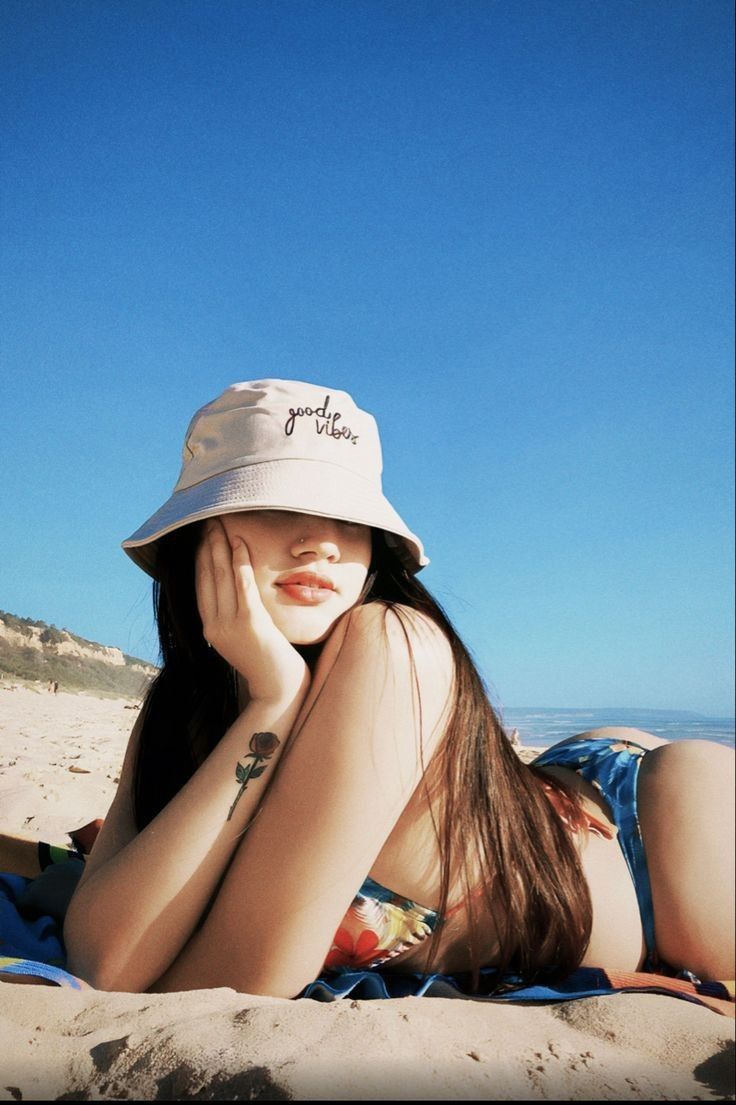 a woman laying on top of a sandy beach next to the ocean wearing a hat