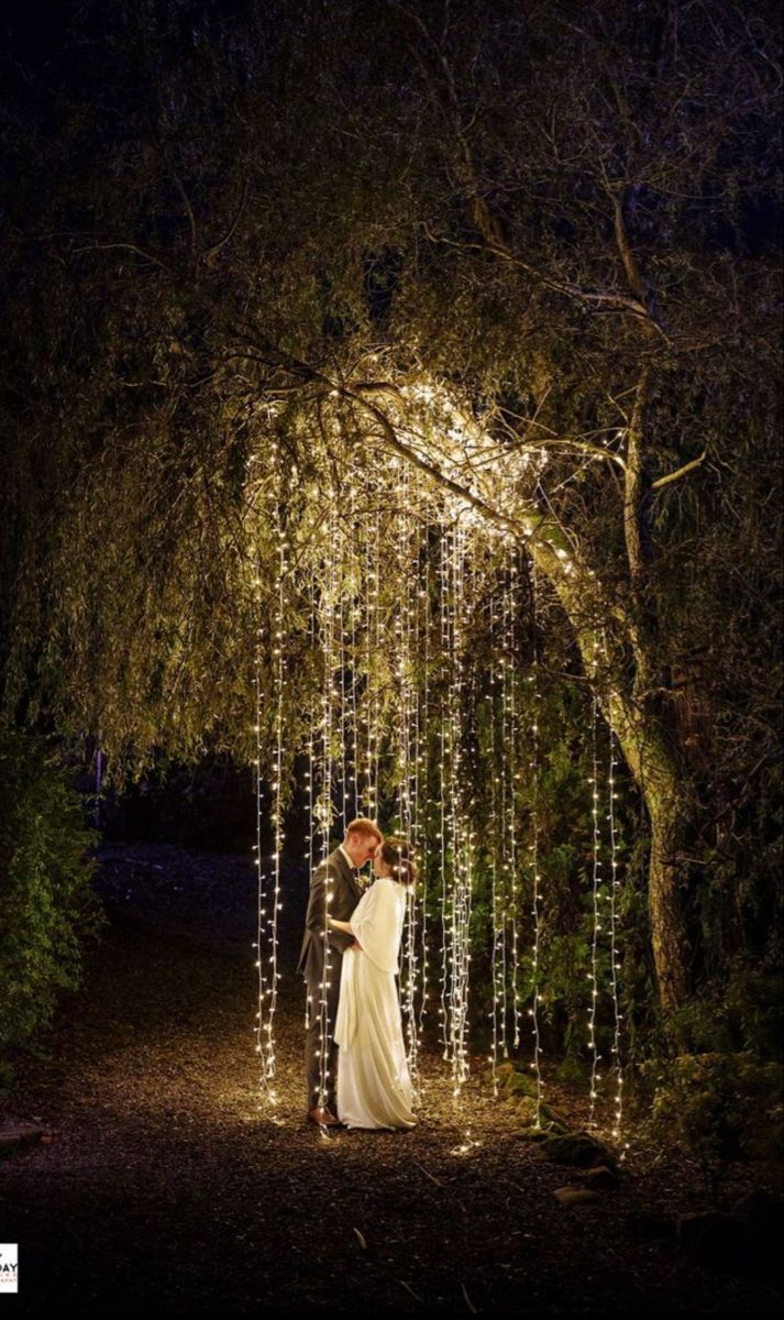 a bride and groom standing under an archway covered with lights at night in the woods