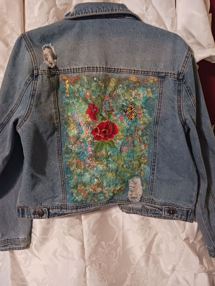 a jean jacket with flowers on it is laying on a white bed sheet and there is a red rose embroidered on the back