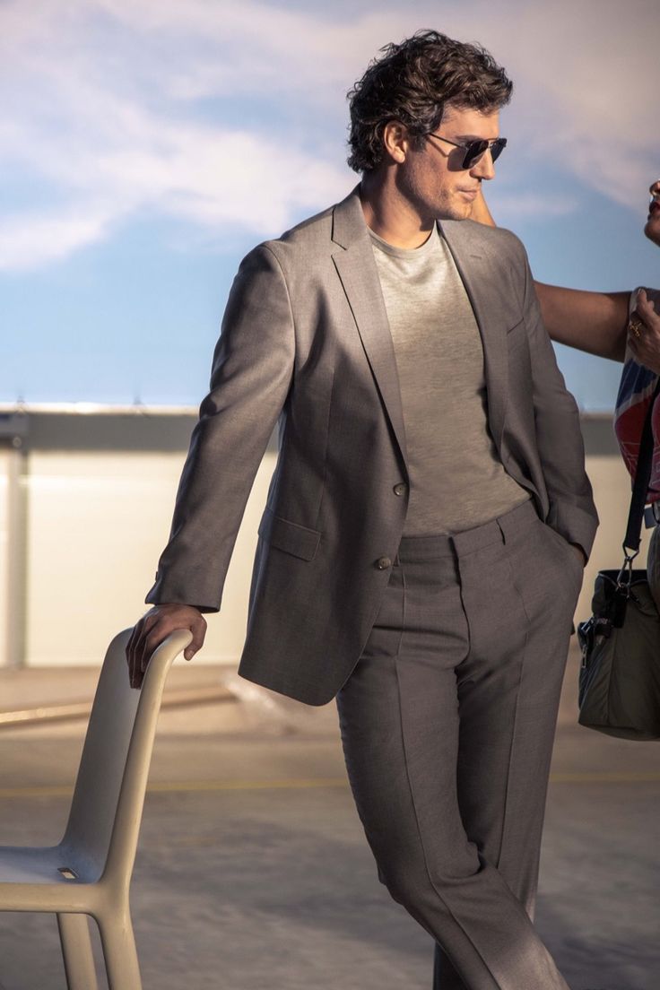 a man in a gray suit and sunglasses is leaning against a chair with his hand on his hip