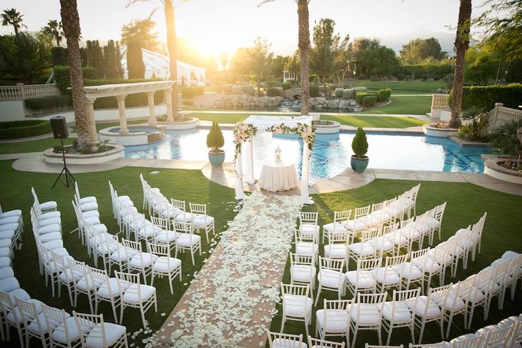 an outdoor ceremony setup with white chairs and flowers on the aisle, in front of a pool