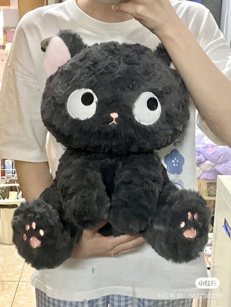 a person holding a black teddy bear with big eyes on it's chest in front of a store display