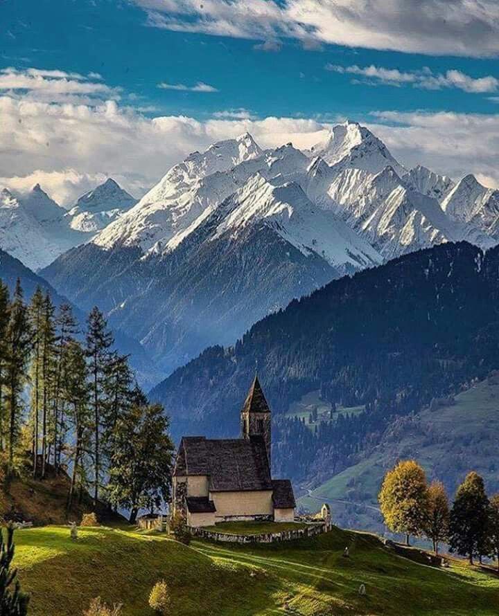 a church in the middle of a mountain range with snow capped mountains in the background