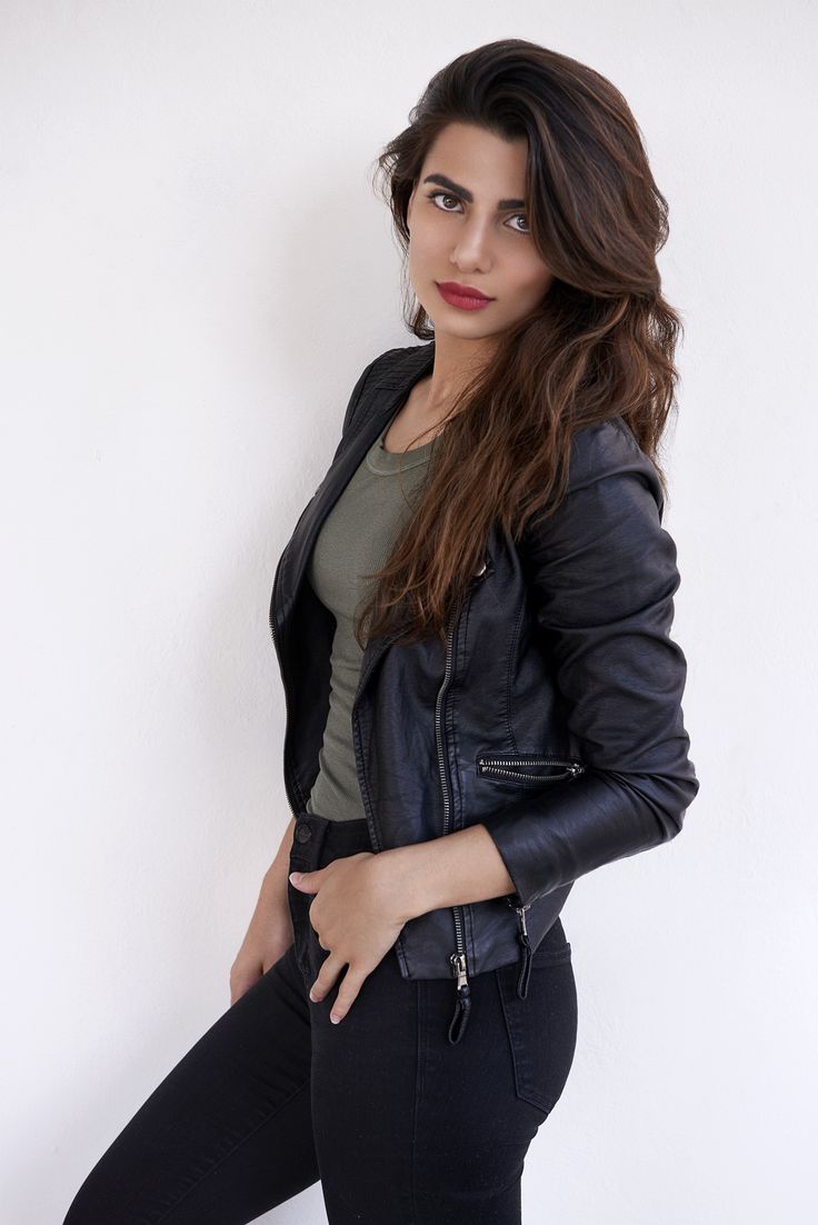 a woman in black leather jacket leaning against a wall with her hands on her hips