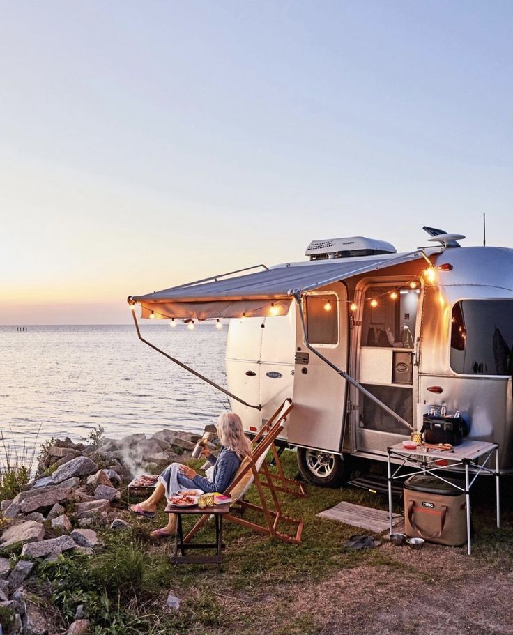 a woman sitting in a chair next to a camper on the shore at sunset