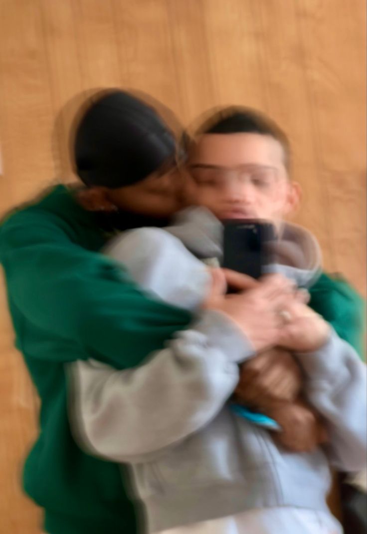 two people hugging each other in front of a wooden wall and one person holding a cell phone