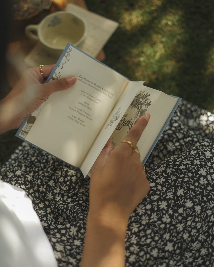 a woman is holding an open book in her hands while sitting on the grass outside