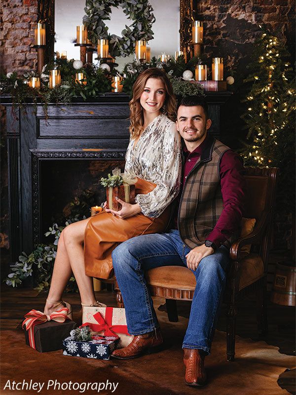 a man and woman sitting on a chair in front of a fireplace with christmas decorations